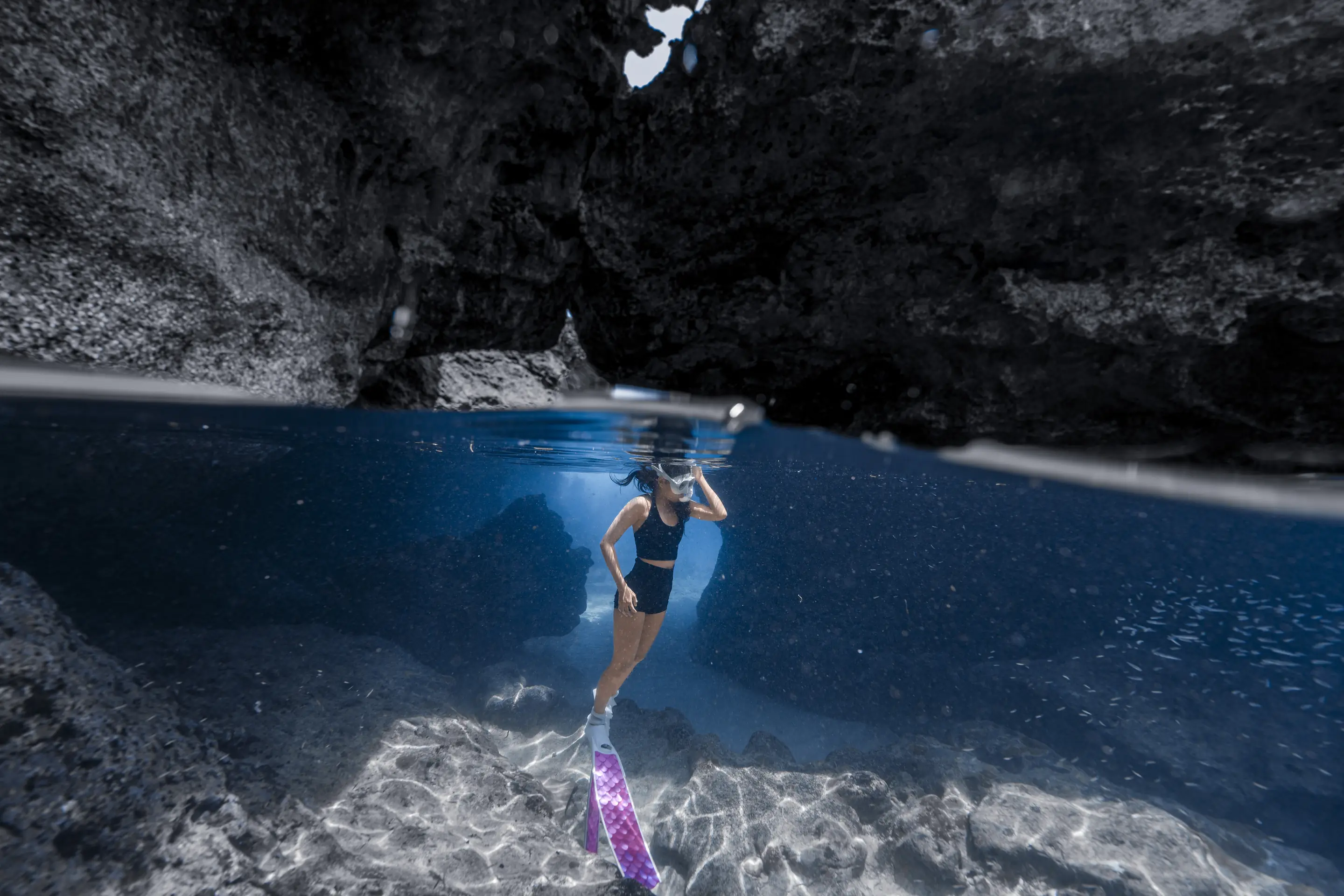 Split-shot di gua laut: freediver dengan fin ungu di bawah permukaan, langit-langit gua di atas air